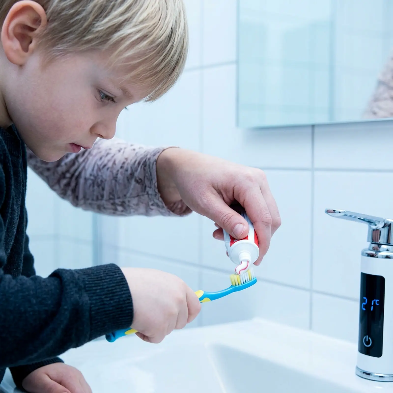 Parent helping a toddler put toothpaste on a toothbrush.