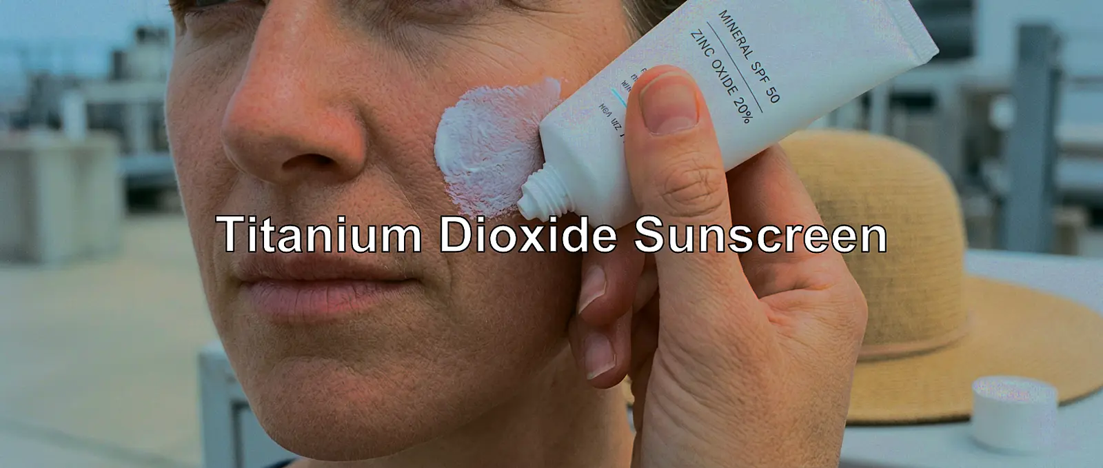 Close-up of a person applying white mineral sunscreen to the cheek; tube and hat visible